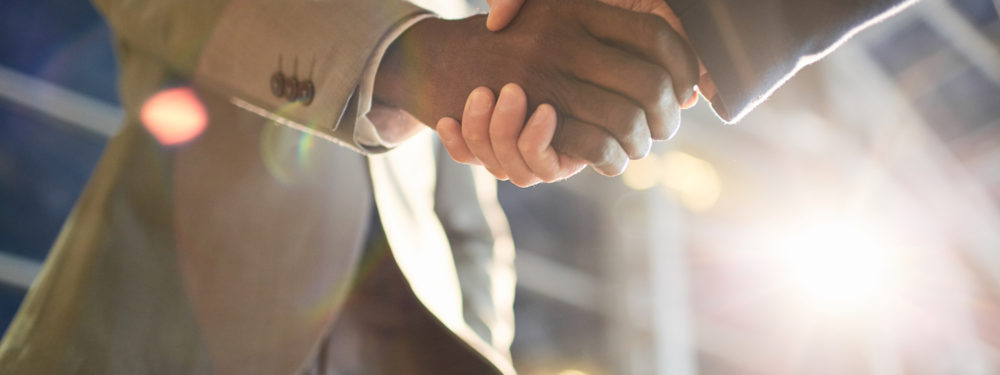 Low angle closeup shot of two business partners in handshake: unrecognizable African -American businessman shaking hands with Caucasian colleague in hall of modern glass office building at night time